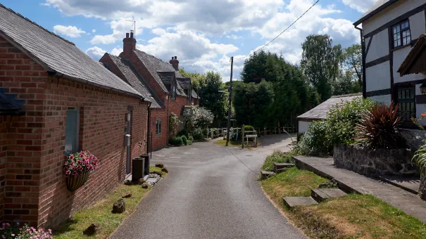 A photograph of a partially cloudy day, a short road trails off with a few houses either side. Tree's line the end where the road veers off right.